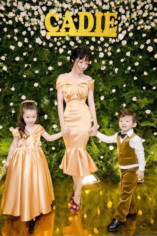 Trong lễ sinh nhật của Candie Mộc Trà, Elly Trần lộng lẫy như công chúa trong bộ váy vàng, thiết kế trễ nải, tôn vinh triệt để vòng 2 và 3 một cách khéo léo, thanh lịch mà vẫn đồng điệu với 2 người con