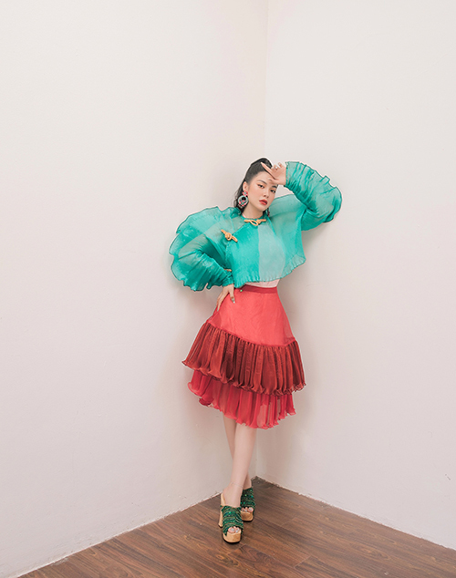 Áo tay phồng lấy cảm hứng từ thân trên của áo dài truyền thống được Liy Chen kết hợp với váy ngắn xếp tầng hiện đại. Nữ diễn viên thể hiện khả năng mix đồ táo bạo khi phối áo xanh bạc hà với chân váy đen và guốc mộc xanh lá cây đậm.